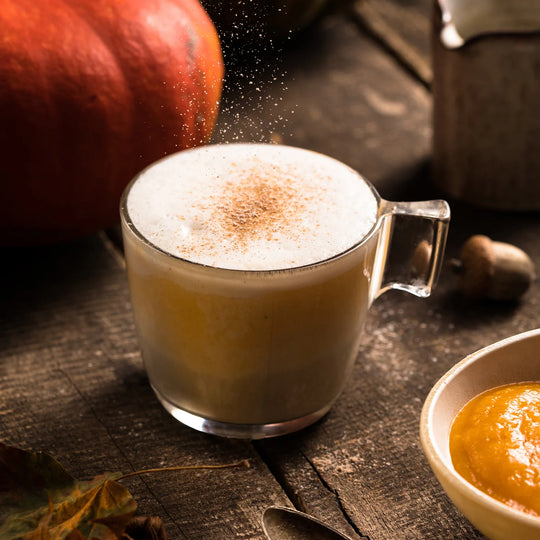 Glass mug of pumpkin spice latte with cinnamon being sprinkled on top, surrounded by pumpkins and a bowl of pumpkin puree.